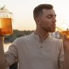 man drinking a glass of kombucha through a straw while holding a jar of kombucha in his other hand