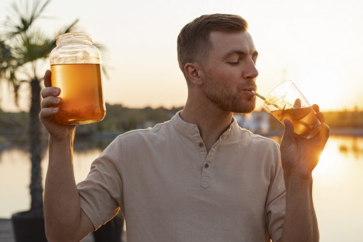 man drinking a glass of kombucha through a straw while holding a jar of kombucha in his other hand
