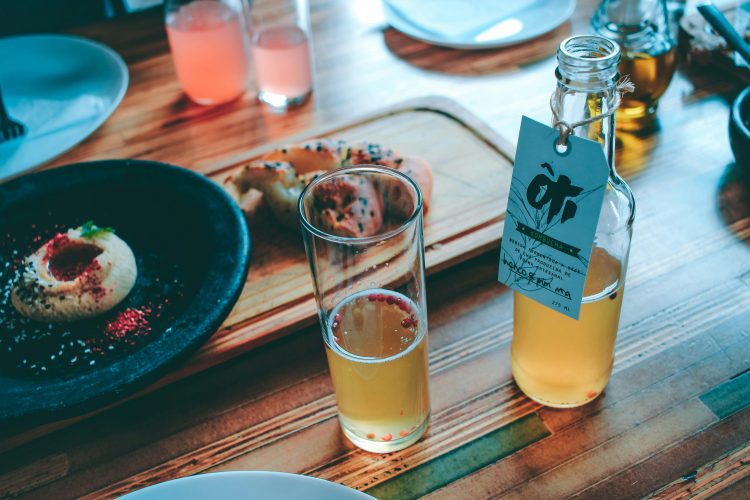 open bottle of kombucha next to a half-filled glass, with food in the background